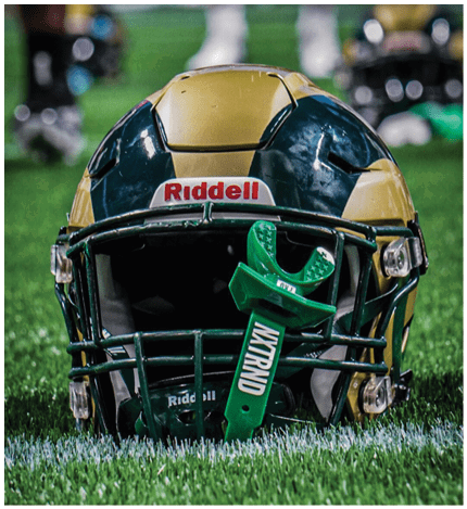 A green and yellow football helmet with a green mouthguard attached is resting on grass with blurred legs and shoes in the background. Description generated by AI