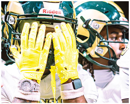 A person wearing yellow gloves, a white sports jersey with gold and green lettering, a watch, and a smartwatch is raising their hands while standing next to others in similar sports uniforms. Description generated by AI