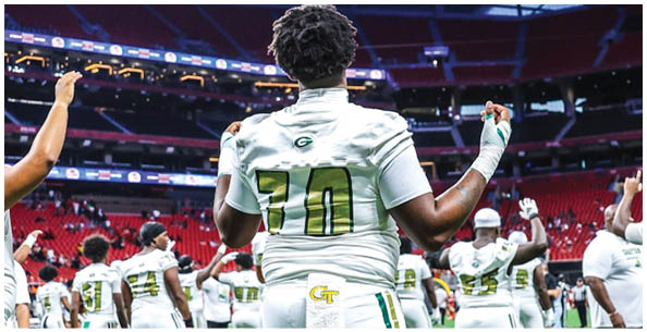 A group of people wearing white sports uniforms with green and yellow accents are standing on a field inside a stadium, with one person in the foreground flexing their arms. Description generated by AI