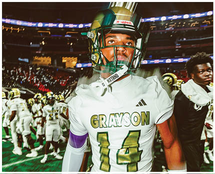 A person wearing a white football uniform with the name Grayson and number 14 stands on a field with other players in the background. Description generated by AI