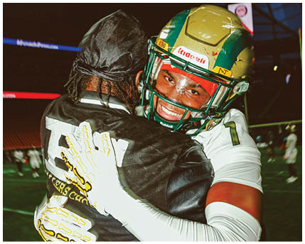 Two people in football gear embrace on a field with one smiling broadly while wearing a helmet. Description generated by AI