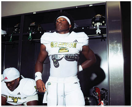 Two people in white football uniforms with “GRAYSON" on the jerseys are in a locker room, one standing confidently and the other sitting and looking down. Description generated by AI