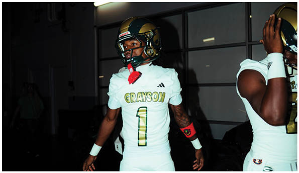 Two people wearing white football uniforms with gold helmets stand indoors, with one person facing forward and the other partially visible on the right. Description generated by AI