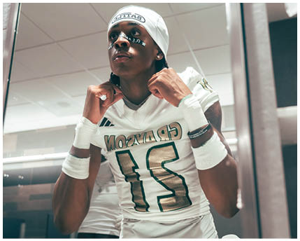 A person wearing a white sports uniform with the number 21 and a white headband is adjusting their collar while sitting in a locker room. Description generated by AI