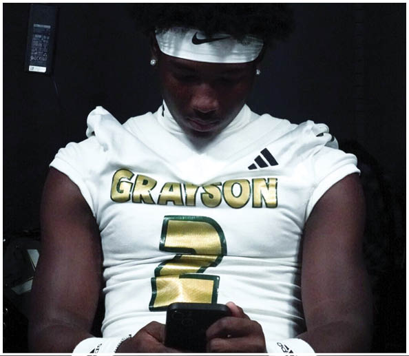 A person wearing a white sports jersey with the word “GRAYSON" and the number 2 in gold is looking down at a phone while seated. Description generated by AI