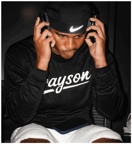 A person wearing a black long-sleeve shirt with the word “Grayson" and white shorts is holding headphones to their ears while looking down at a phone resting on their lap. Description generated by AI