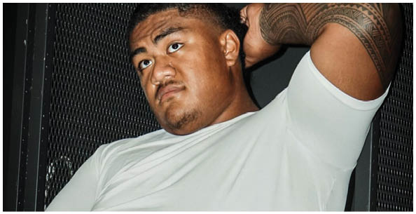 A person wearing a white t-shirt is sitting against a dark mesh background with one arm raised. Description generated by AI
