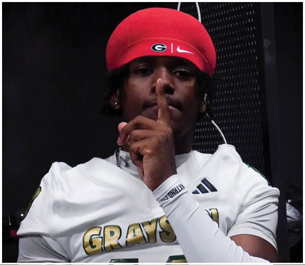 A person wearing a white football jersey with the word “GRAYS" and the number 18 is holding a gold and green helmet while sitting. Description generated by AI