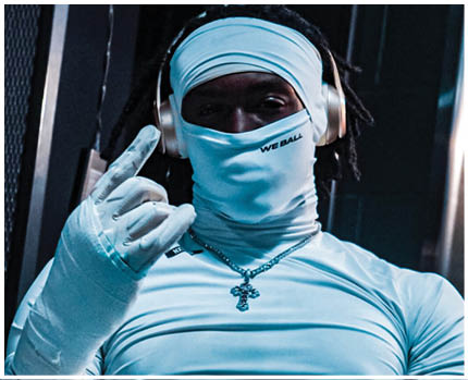 A person wearing a tight light blue shirt, headphones, a necklace with a cross pendant, and a matching light blue face covering and glove is making a hand gesture. Description generated by AI
