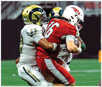 Two people in football uniforms are engaged in a tackle on a green field, with one holding the ball and the other wrapping arms around them. Description generated by AI