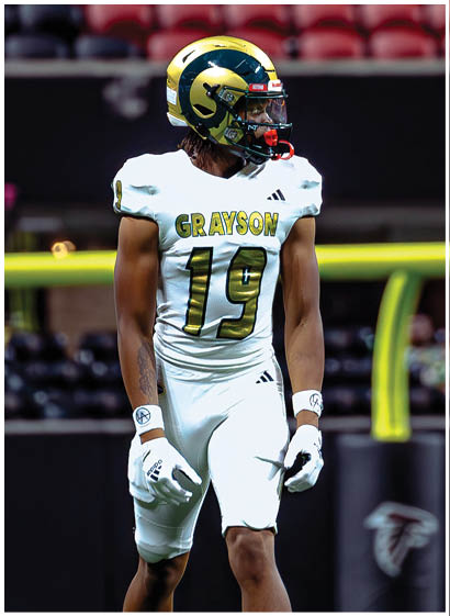 A person wearing a white football uniform with the name “Grayson" and number 19 stands on a football field near a goalpost. Description generated by AI