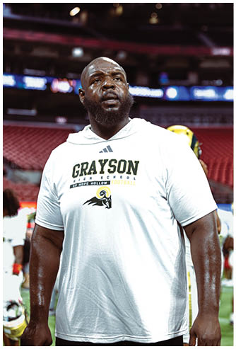 A person wearing a white Grayson High School football hoodie stands in an indoor sports arena with blurred seating and lights in the background. Description generated by AI