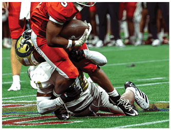 A person in a red football uniform is holding a football while being tackled by a person in a white and gold football uniform on a green field. Description generated by AI