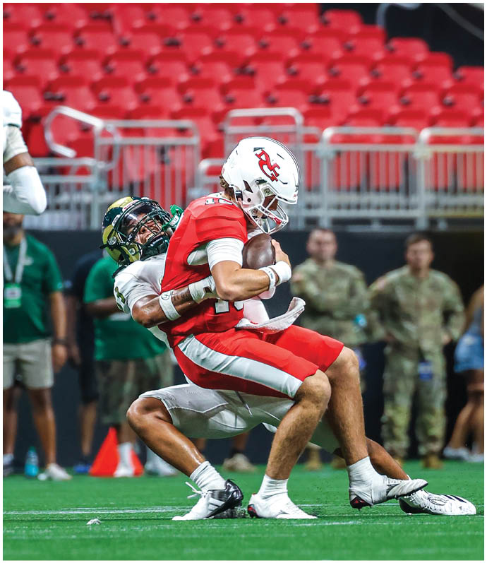 A person in a red football uniform is holding a football while being tackled by another person in a white uniform on a green field with red stadium seats in the background. Description generated by AI