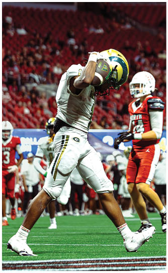 A person in white football gear is celebrating on the field while holding a football near their helmet with other players in red uniforms in the background. Description generated by AI