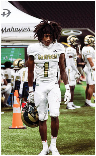 A person in a white football uniform with “GRAYSON" and the number 1 on the jersey is standing on a field holding a helmet, with other players and equipment visible in the background. Description generated by AI