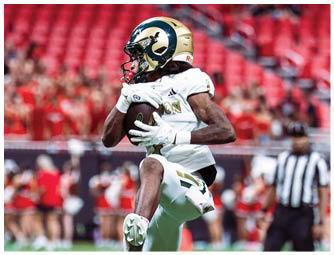 A person in a white football uniform with a gold and blue helmet is catching a football on a field with a blurred crowd and referee in the background. Description generated by AI