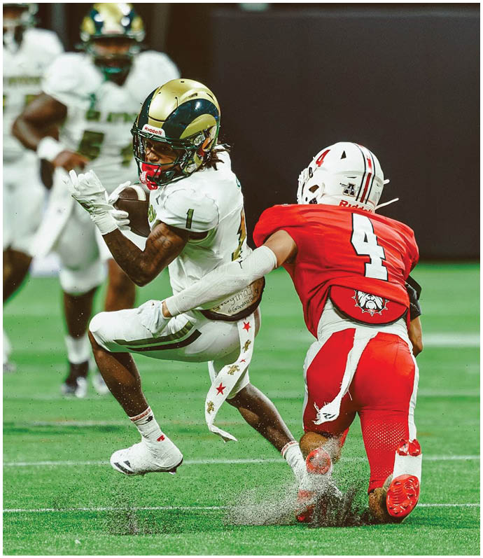 A person in a white and gold football uniform is running with the ball while another person in a red and white football uniform attempts to tackle them on a green field. Description generated by AI