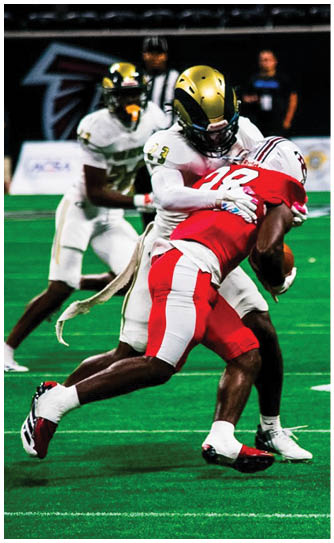 Two people in football uniforms are engaged in a tackle on a green field with others and photographers visible in the background. Description generated by AI