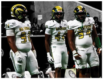 Three people wearing football uniforms and cleats stand on a field with a blurred background. Description generated by AI