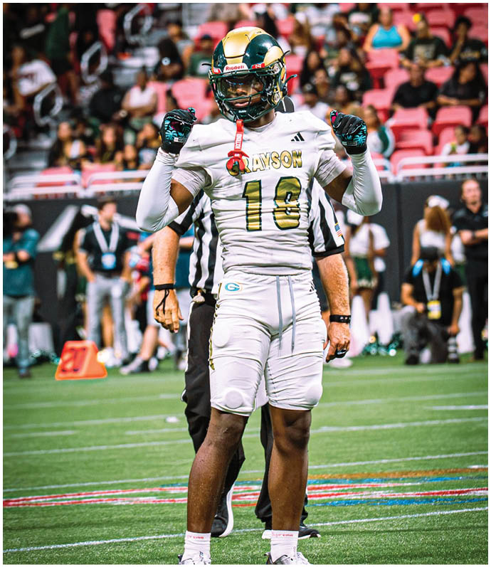 A person wearing a white football uniform with the number 18 and the name Grayson is standing on a football field with arms raised.   Description generated by AI