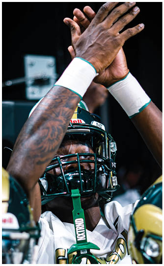 A person wearing a football helmet and jersey raises their tattooed arms above their head in a celebratory gesture during a game. Description generated by AI