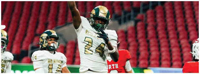 A person in a white and gold football uniform with the number 24 is celebrating on the field while other players in similar and red uniforms stand nearby in a stadium with red seats. Description generated by AI