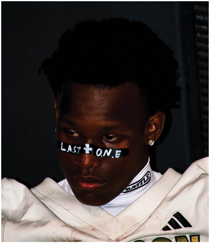 A person with short curly hair wears a white garment and has black tape with the words “LAST ONE" written across the nose. Description generated by AI