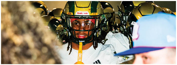 A person wearing a football helmet and a white jersey with the word “GRAYSON" stands among other people, with two blurred individuals in the foreground. Description generated by AI