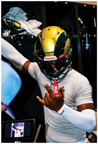 A person wearing a gold and green football helmet and a white shirt is making a hand gesture while standing in a locker room. Description generated by AI