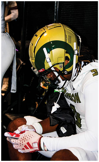 A person wearing a yellow and green football helmet and a white jersey with green lettering is looking downward. Description generated by AI
