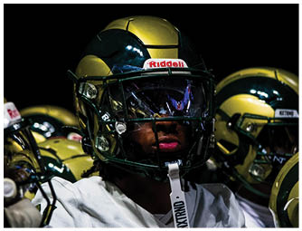 A person wearing a football helmet and white jersey is sitting on a bench with other players in the background. Description generated by AI