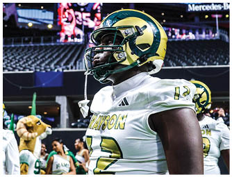 A person wearing a yellow and green football helmet and white jersey is standing in a stadium with empty seats and a large screen in the background. Description generated by AI