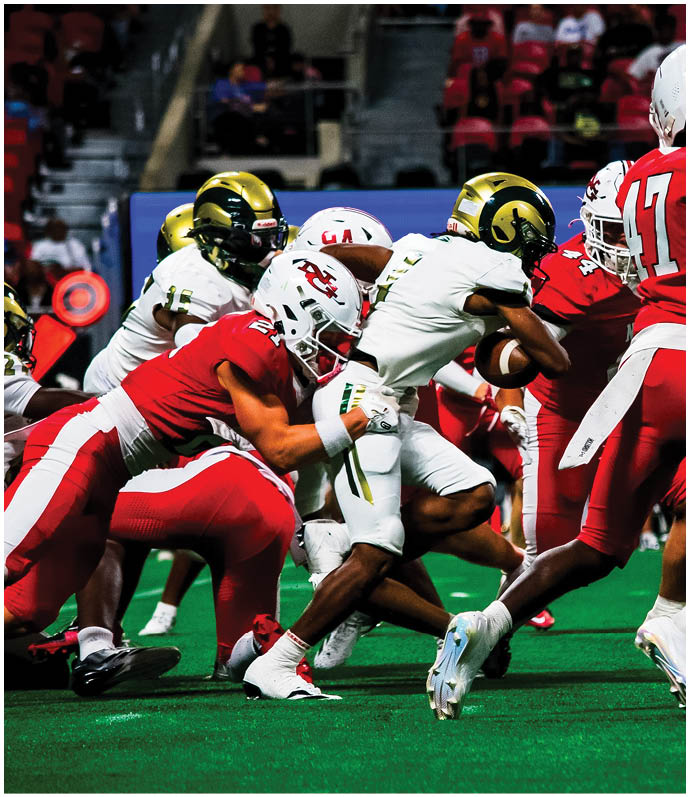 People wearing red and white and others in white and gold uniforms are engaged in a football game on a green field inside a stadium. Description generated by AI
