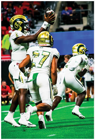 Three people in white and green football uniforms with gold helmets are on a football field, with one person holding a football above their head. Description generated by AI