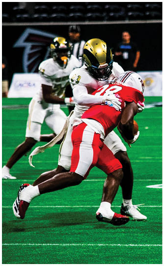 Two people in sports uniforms are engaged in a tackle on a green football field with white yard lines. Description generated by AI