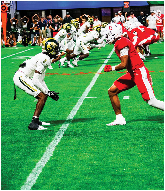 Two opposing American football teams in red and white and white and yellow uniforms are lined up facing each other on a green field, ready for a play. Description generated by AI