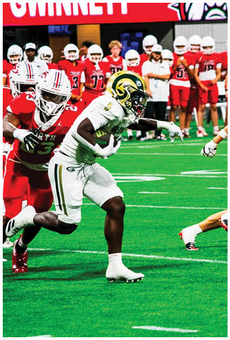 A person in white football gear with a green and yellow helmet is running with the ball while another person in red football gear is attempting to block them on a green field with a crowd in the background. Description generated by AI