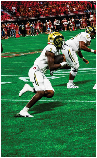 A person in a white football uniform and gold helmet is running on a green field while holding a football, with other players and spectators visible in the background. Description generated by AI