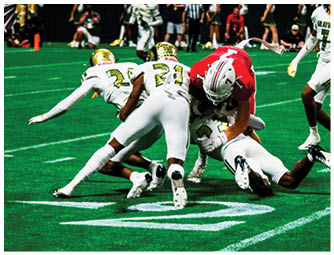 Three people in athletic clothing are diving or sliding on a green sports field near a white yard line marking. Description generated by AI