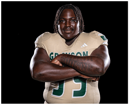 A person with braided hair wearing a beige sports jersey with the word “GRAYSON" and the number 5 is posing with arms crossed, showing a tattoo on one arm against a black background. Description generated by AI