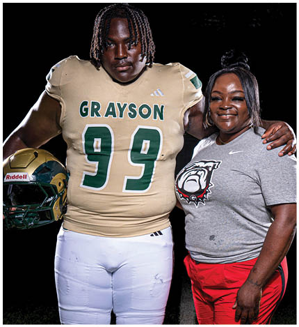 Two adults are posing together with one wearing a football uniform holding a helmet and the other wearing a gray t-shirt and red pants with an arm around the first person. Description generated by AI