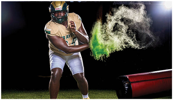 A person in football gear is holding a football while green powder explodes from the ball against a dark background on a turf field. Description generated by AI