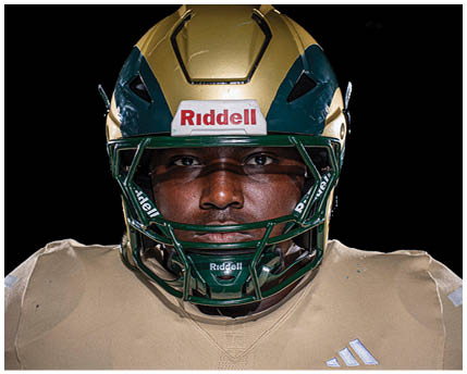 A person wearing a green and gold football helmet and a beige sports jersey is looking directly at the camera. Description generated by AI