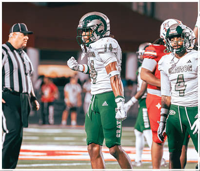 Two people in white and green football uniforms stand on a field with one clenching a fist and a referee and opposing players in red uniforms visible in the background. Description generated by AI