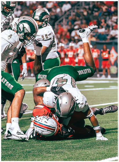 Two football players in green and white uniforms tackle a player in an orange and white uniform on a grassy field during a game. Description generated by AI