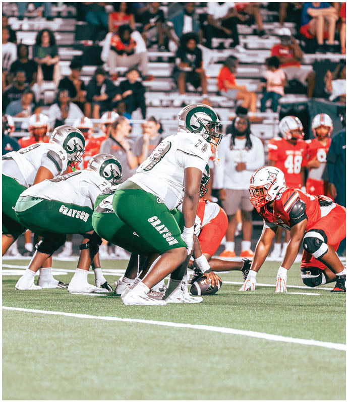 Several people in football uniforms are lined up on a field facing each other, preparing for a play during a game with spectators in the background. Description generated by AI