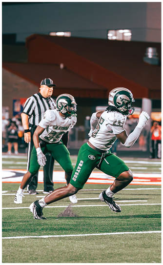 Two football players in green and white uniforms are running on a field with a referee standing behind them. Description generated by AI