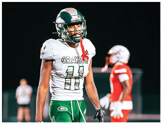 A person wearing green football pants and a white jersey with the number 11 stands on a football field with another person in red football gear in the background. Description generated by AI