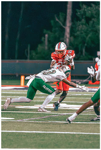 A person in a red football uniform is running with the ball while another person in a white and green uniform lunges forward attempting to tackle them on a football field. Description generated by AI